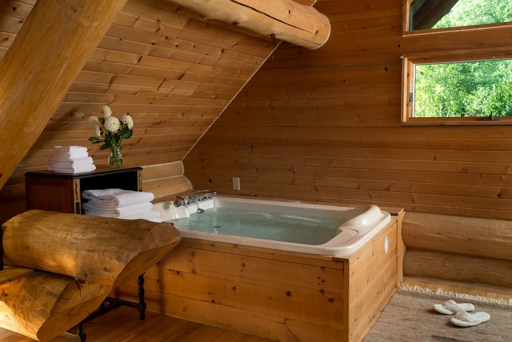 Loft whirlpool bathtub with wood surround, log and wood walls, and window with views into the treetops.