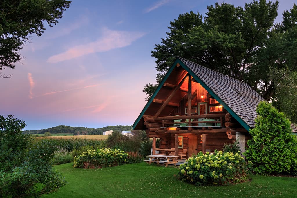 Exterior of Paul Bunyan log cabin with warm lights glowing inside, landscaped shrubs outside, and a pink and blue sky at dusk above.