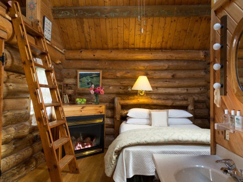 Cozy interior of cabin with natural color log walls, car siding ceiling, queen bed, corner fireplace and wooden ladder to loft.