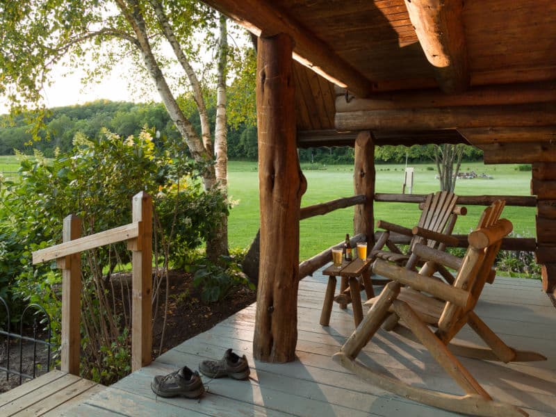 Open porch with log rocking chairs, steps with hand rail and edged with shrubbery and birch tree, and views looking over bright green lawn.