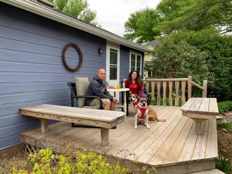 A man and a woman enjoy a lovely day with two dogs at their feet while sitting on patio furniture on a wooden deck outside a Wedgewood blue cottage.