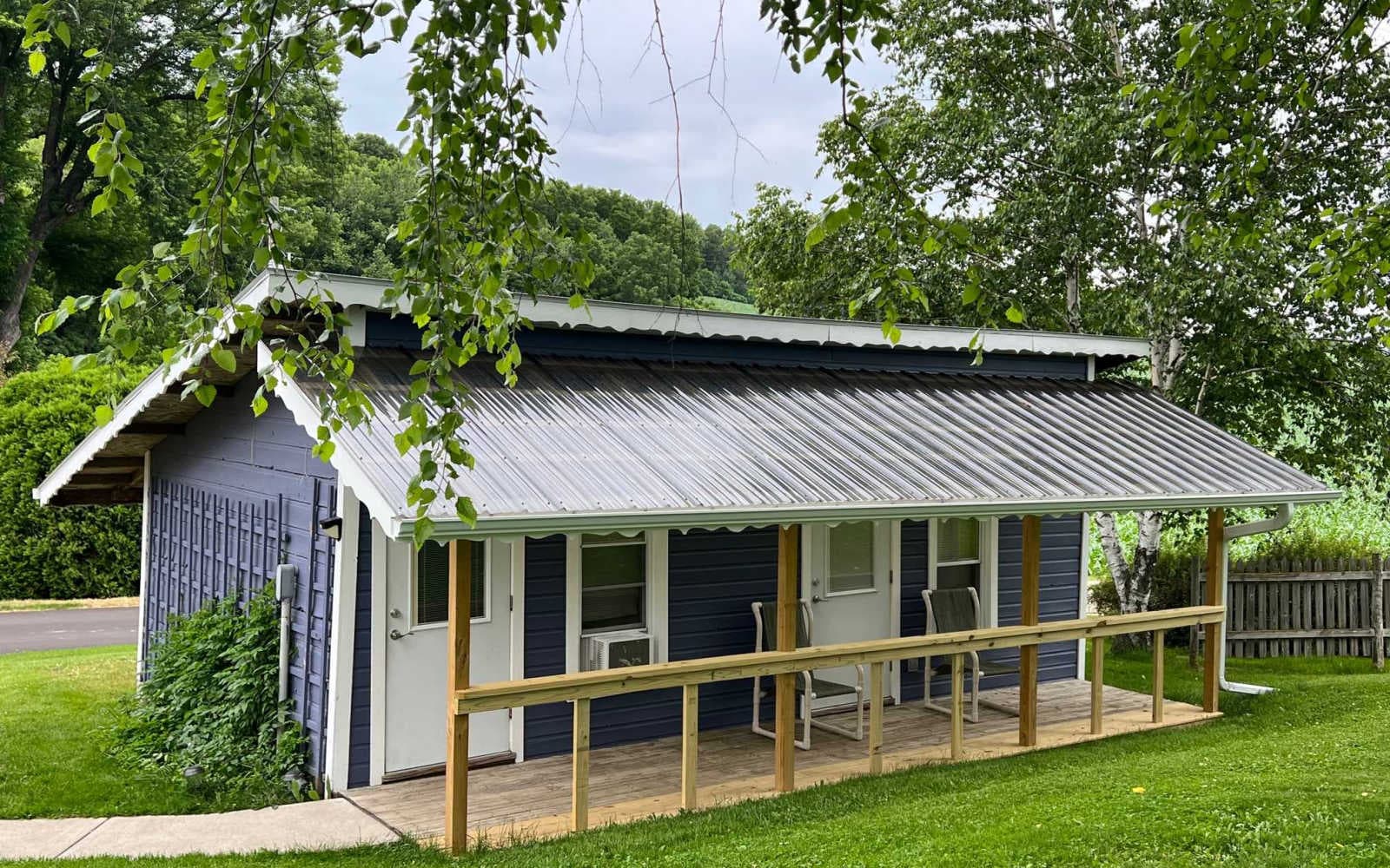 The Camping Cabin has blue exterior with white trim and doors, a front patio with chairs, and a metal roof.