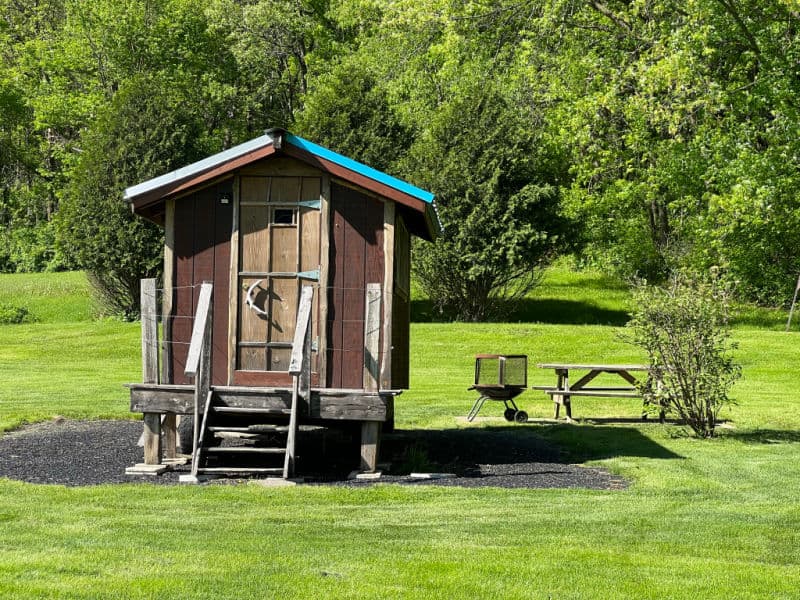 Snuggalo is a wooden tiny house with wooden stairs with handrails leading to the front door.  It is located near the woods with a picnic table and portable firepit.