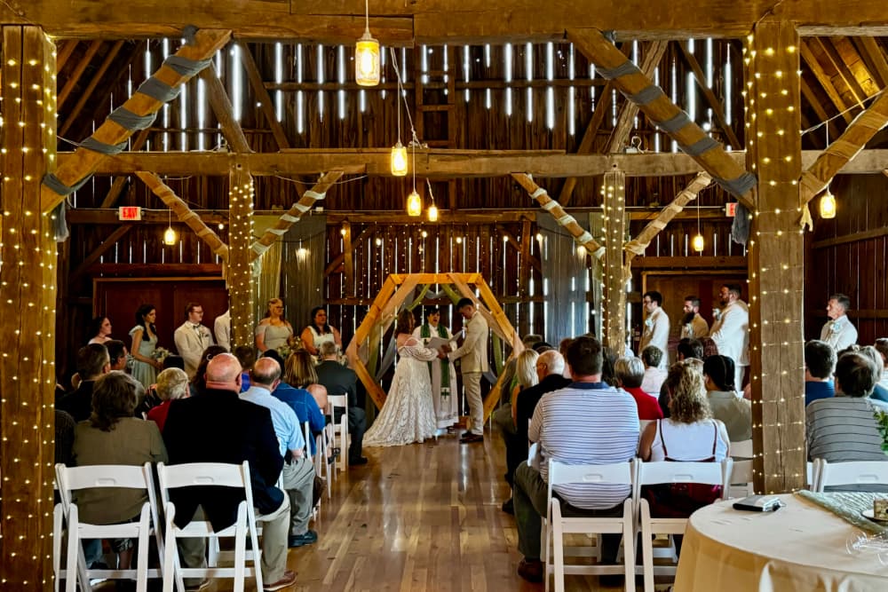 A wedding couple saying their vows inside an elegant barn with shiny wood floors, wood beams covered with lights, and guests sitting in chairs watching the wedding couple.