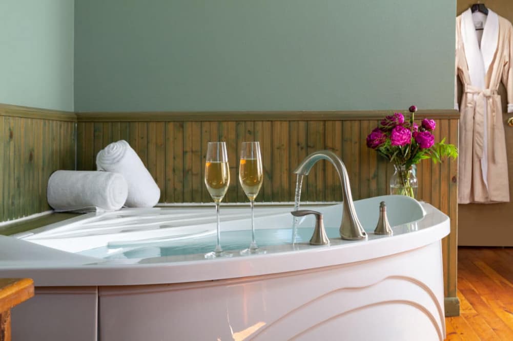 A large corner jetted tub filled with water, 2 glasses of champagne on the edge,2 rolled towels on the corner of the tub, a vase of pink flowers nearby, along with robes hanging on hooks.