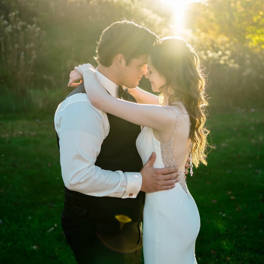 A bride and groom dressed in wedding attire outside embracing with the sun shining through trees behind them.