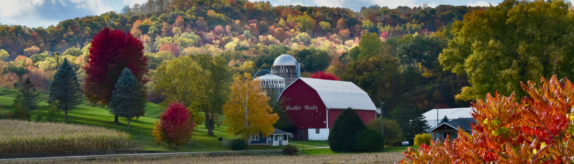 Red barn, silos and other farm buildings in the fall with fall color trees, green grass and blue sky.