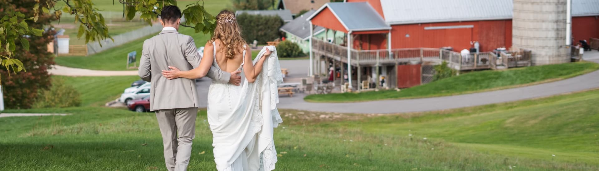 A couple dressed in wedding attire walking through green grass towards a large red barn.