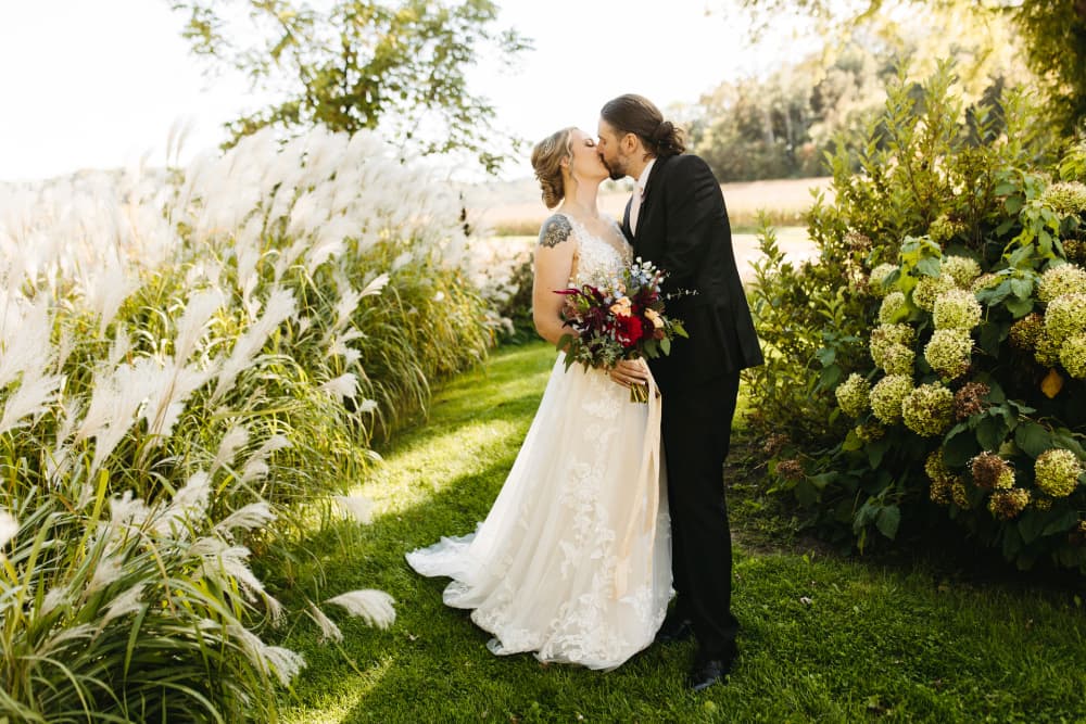 A bride and groom kissing in an outdoor field with hydrangeas and wheat grass.