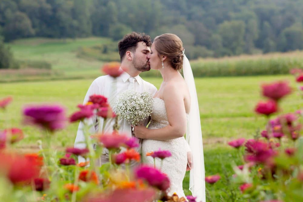 A bride and groom dressed in wedding attire, the bride holding a bouquet of fresh baby's breath, kissing in the midst of pink and orange flowers in the garden, green grass and woods in the background.