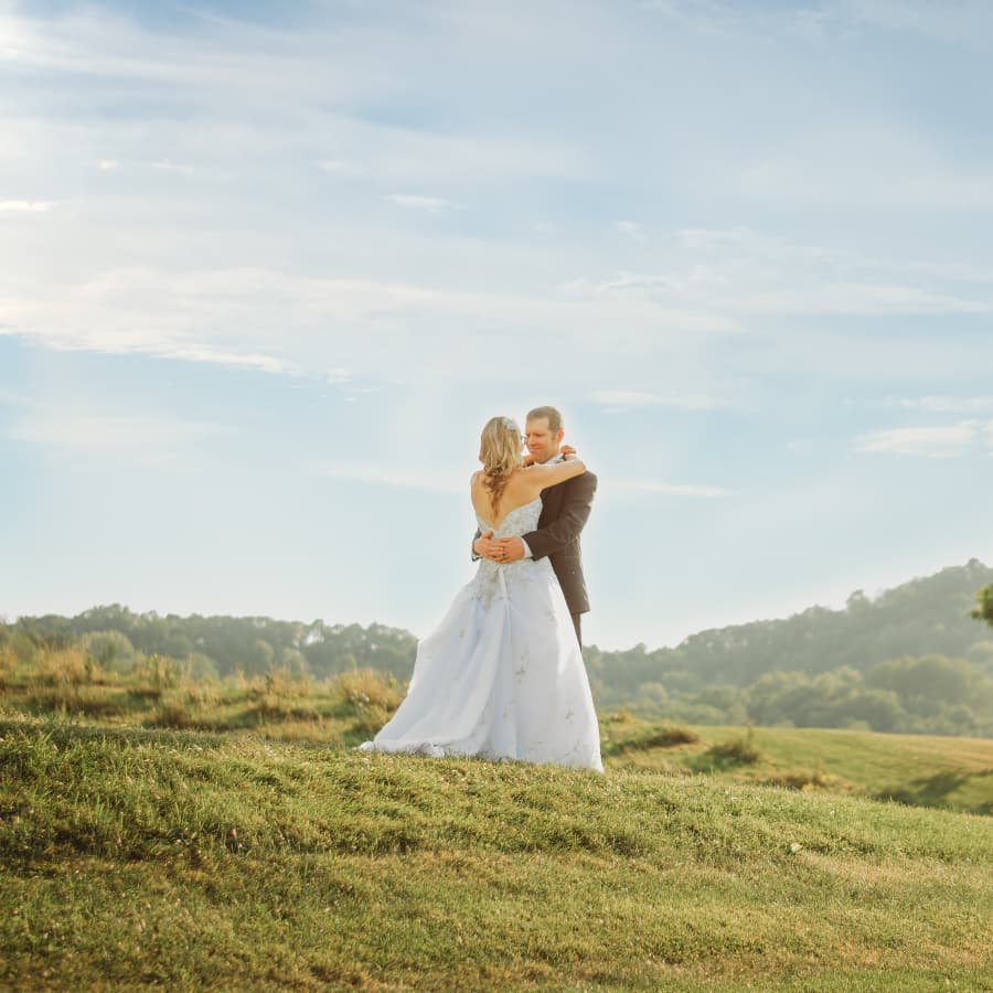 Groom and bride in wedding clothes standing on green hill with blue skies in background