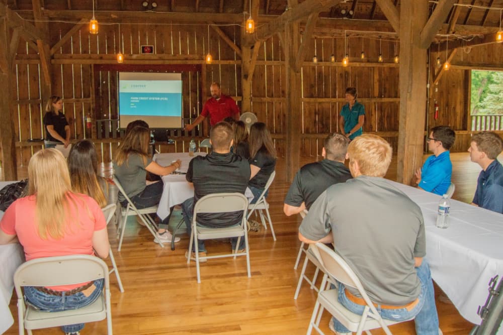 A business meeting with several tables and chairs inside a large barn and people watching someone give a presentation.