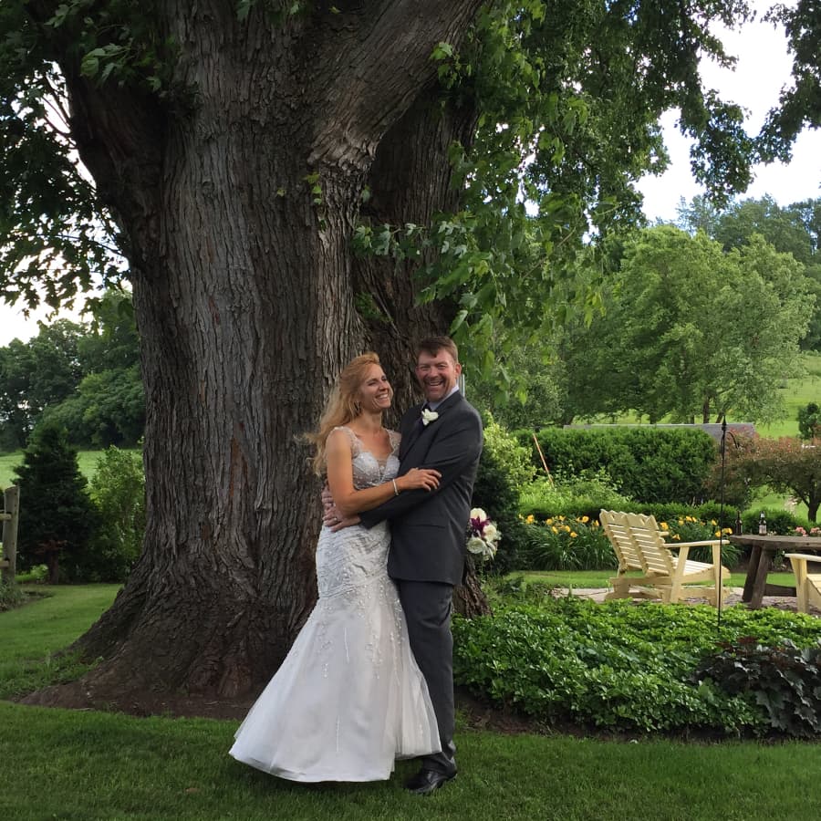 A smiling couple dressed in wedding attire in front of a large oak tree, with a wood table and chairs nearby.