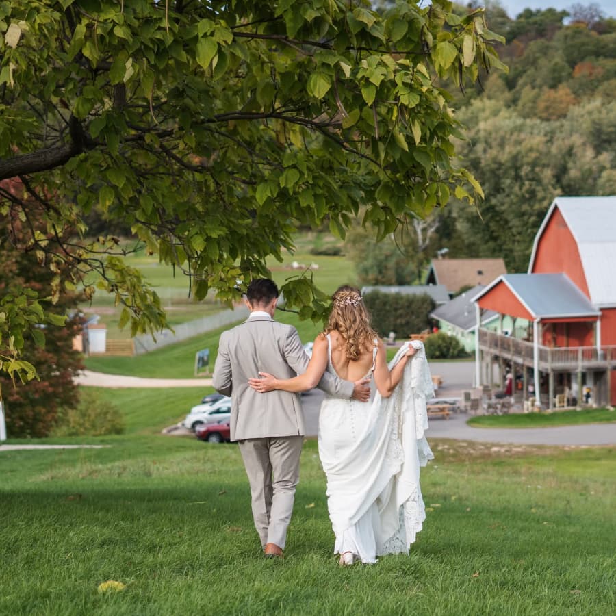 A couple dressed in wedding attire walking through green grass towards a large red barn.