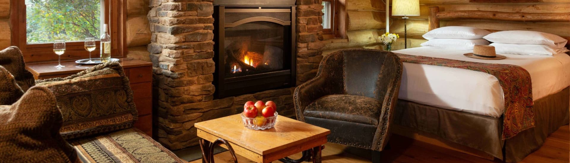 A room in a log cabin with a bed, fireplace, sofa and coffee table with a bowl of fresh apples, and a nearby table in front of a window with a bottle of white wine and 2 glasses filled with wine.