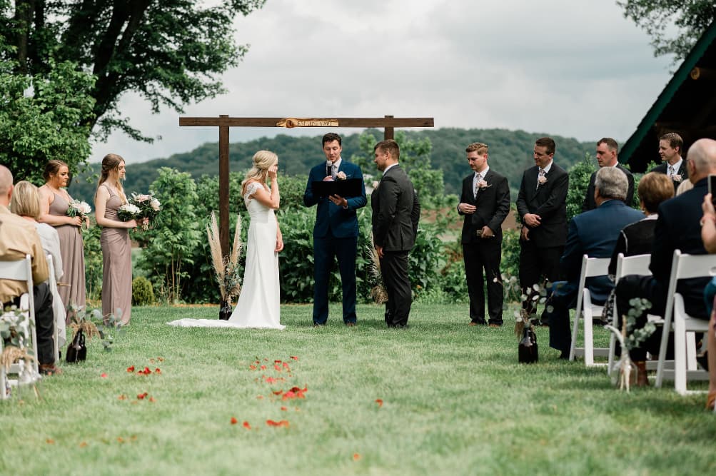 A bride and groom saying their vows in an outdoor ceremony amongst wedding guests on either side sitting in chairs, and rolling hills and woods behind them.