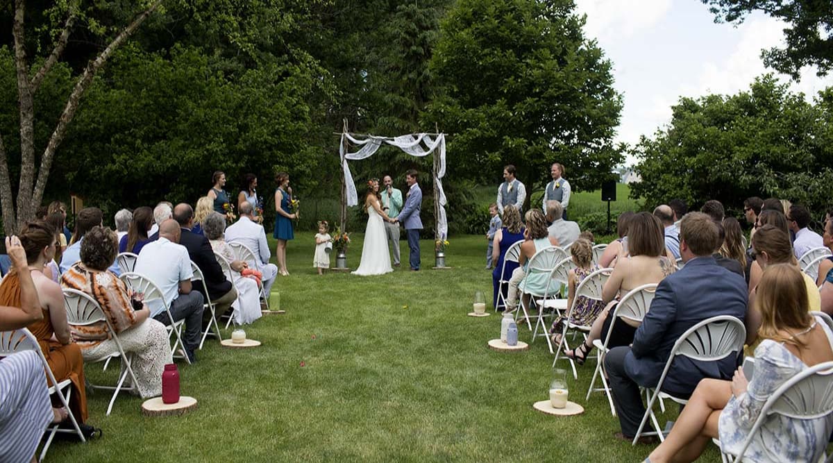 A couple saying their vows in an outdoor wedding setting with a wood arch with draped fabric above them, and guests looking on.