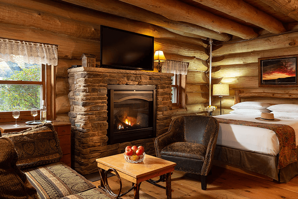 Stone fireplace log walls and bed with white linens, tv, comfortable chair and table with wine glasses