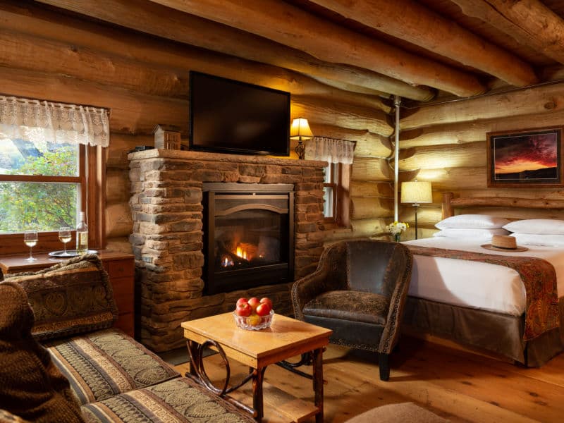 Cozy interior of log cabin with natural log walls, queen bed, and sitting area near gas fireplace with stone surround and flat screen TV above.