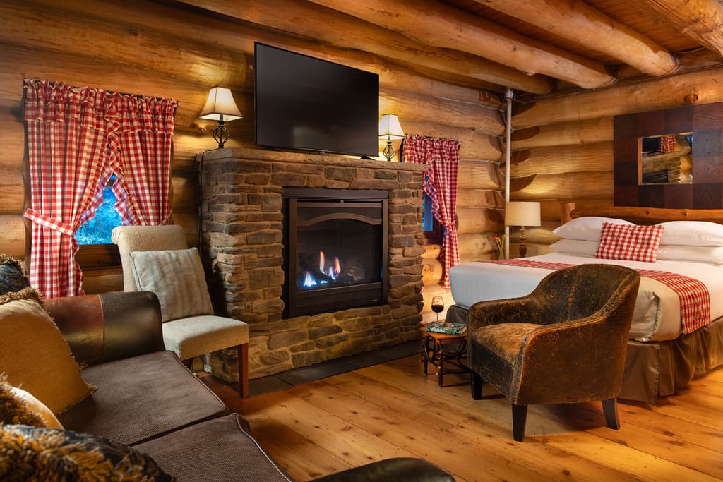 Cozy interior of log cabin with natural log walls, sofa, queen bed, and sitting area near gas fireplace with stone surround and flat screen TV above.