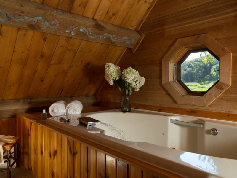 Whirlpool bathtub in loft with wooden surround, glass vase with flowers, and an octagonal window with treeline and lawn view.