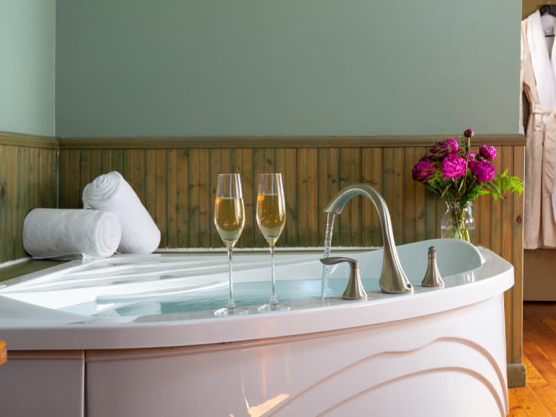 Whirlpool tub with two glass champagne flutes set on the ledge, along with plush white towels rolled up and a glass vase with magenta roses.
