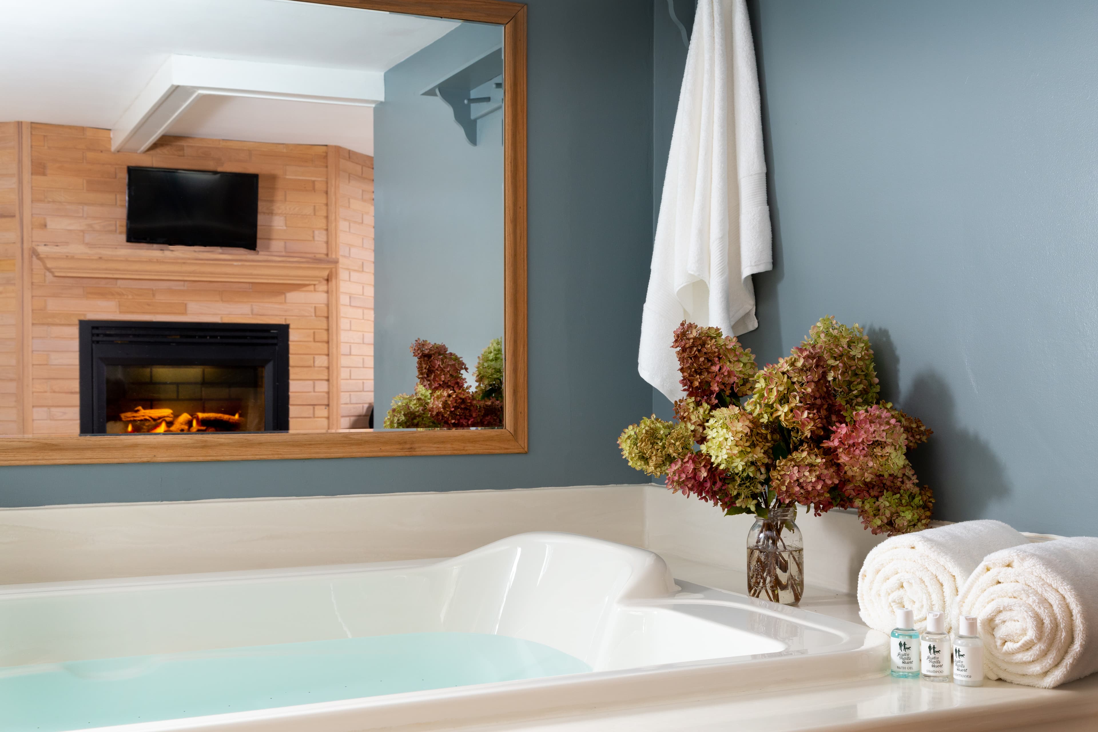 Very large double BainUltra whirlpool bathtub with white surround and mirror on wall above showing a reflection of a fireplace with oak brick surround and flat screen TV above.