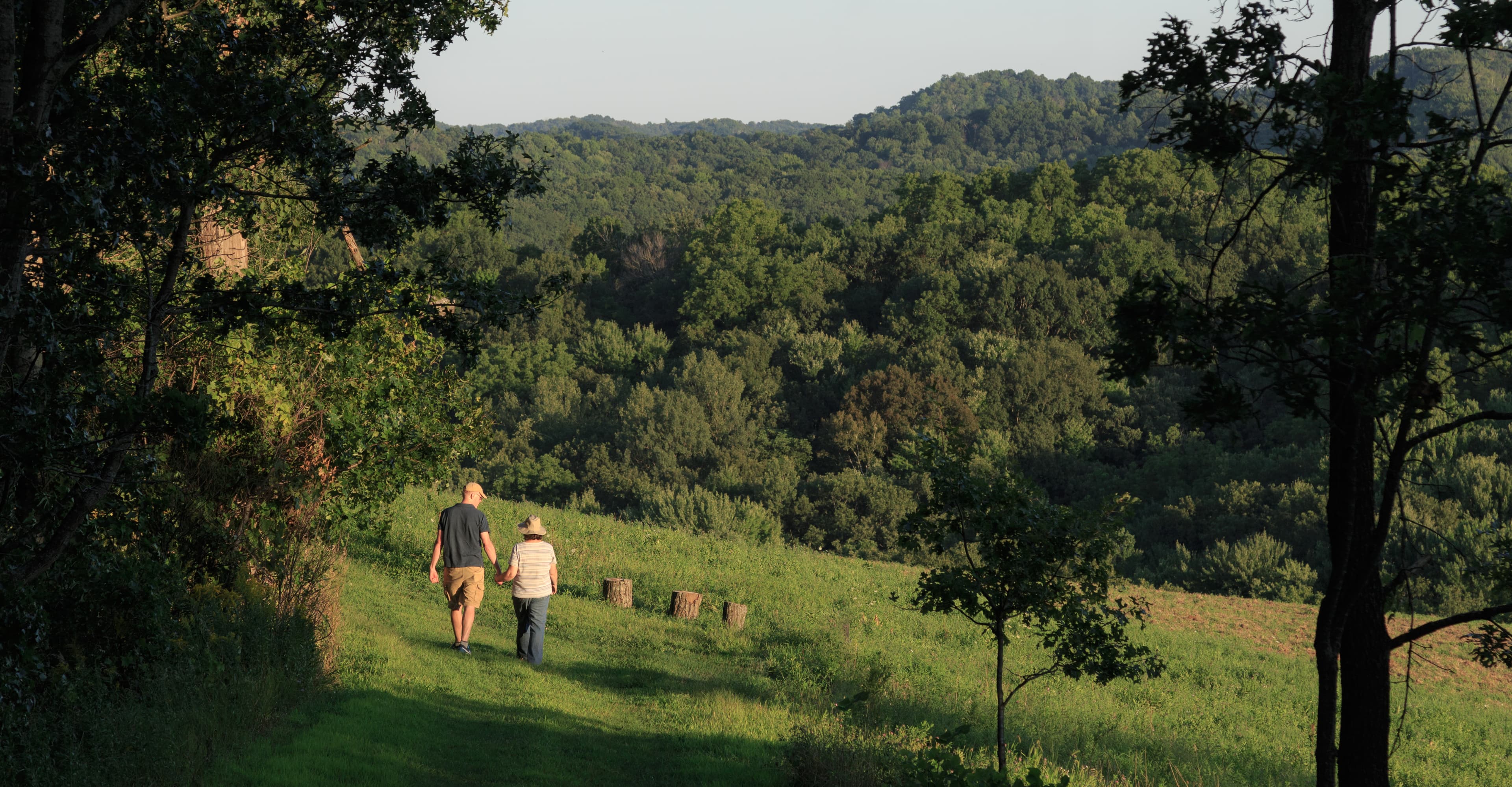 Two people walking on trail with field and woods in background.