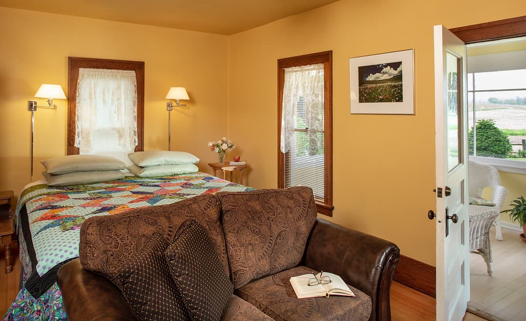 Warm and inviting bedroom with gold walls, Select Comfort grand king bed topped with a quilt and a cozy loveseat at the foot.  A door stands open leading to the east porch with wicker furniture.