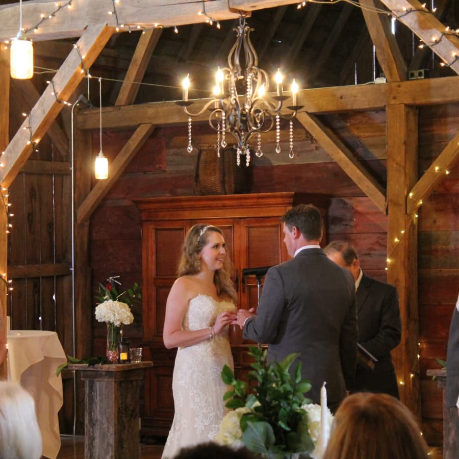 A couple dressed in wedding attire in an elegantly decorated barn, exchanging rings in front of an officiant.