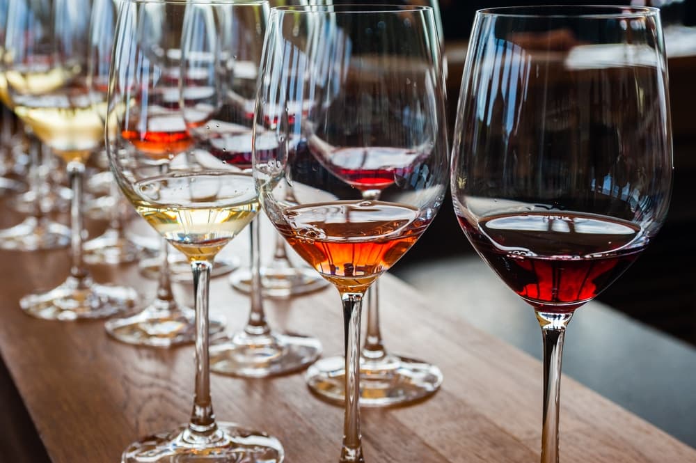 A row of wine glasses filled with various colors of wine on a wooden surface.