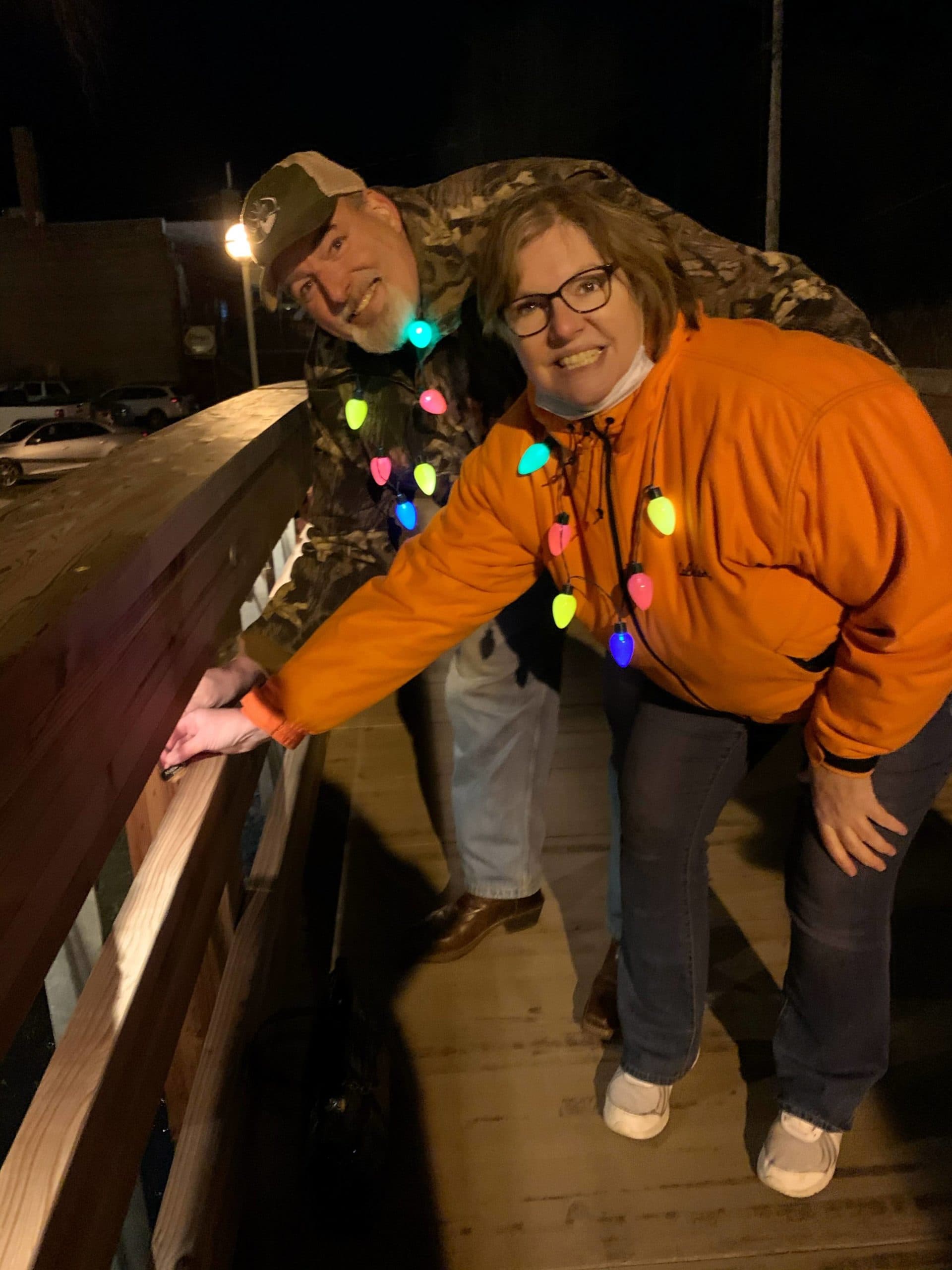 Two people happily posing together while decorating a railing with colorful lights at night.
