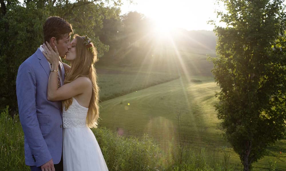 A couple dressed in wedding attire sharing a kiss outside with the sun shining through the trees behind them.