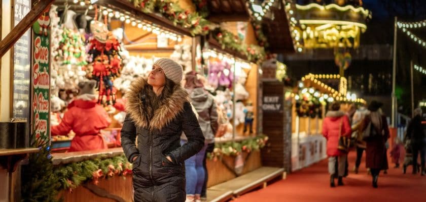 A woman in a fur-trimmed jacket gazes at festive market stalls adorned with lights and decorations.