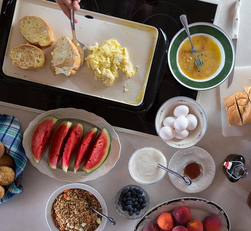 An aerial view of a breakfast table with scrambled eggs, hard boiled eggs, fresh watermelon slices, granola, yogurt, and bowls of fresh peaches and blueberries