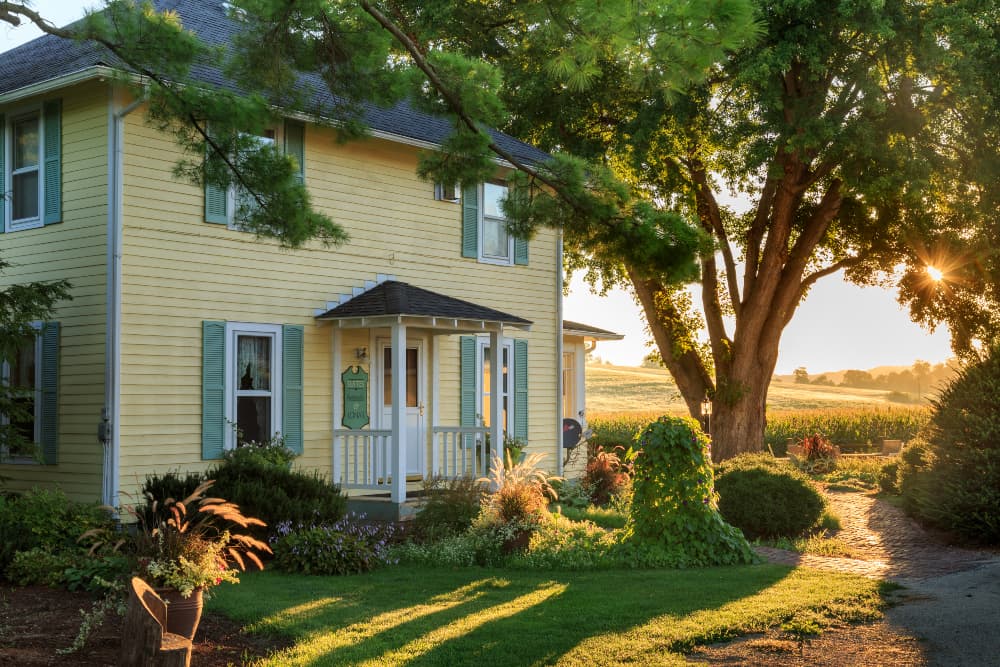 Yellow 2 story sided house, with light green shutters, and small portico with large tree in front.