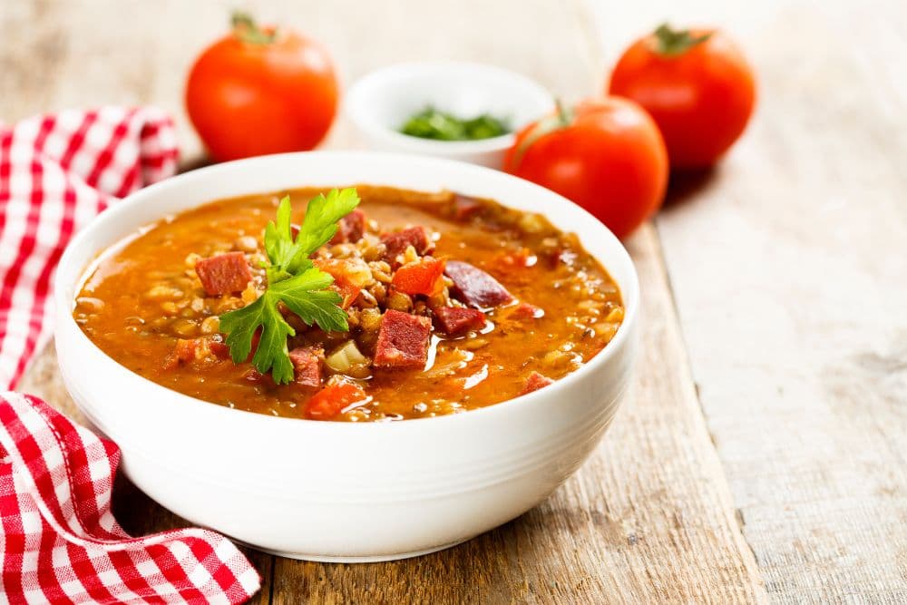 A white bowl filled with a hearty homemade beef and barley soup, whole tomatoes and a bowl of fresh parsley next to it, along with a red and white checkered cloth napkin.
