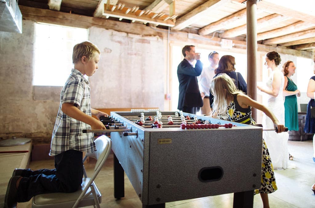 Two children play foosball in a basement while adults socialize in the background.