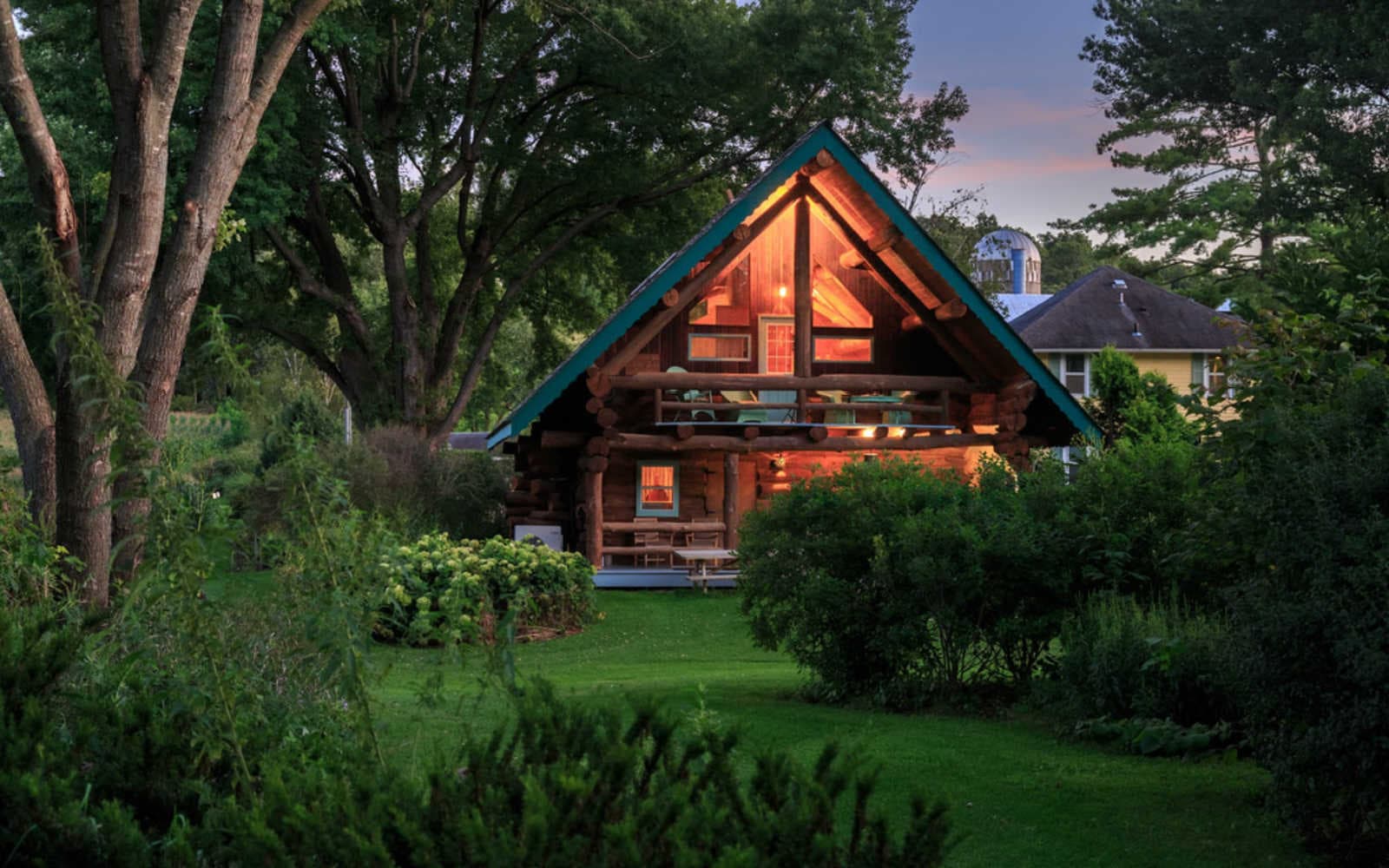 A cozy log cabin with a green roof illuminated at dusk, surrounded by lush greenery.
