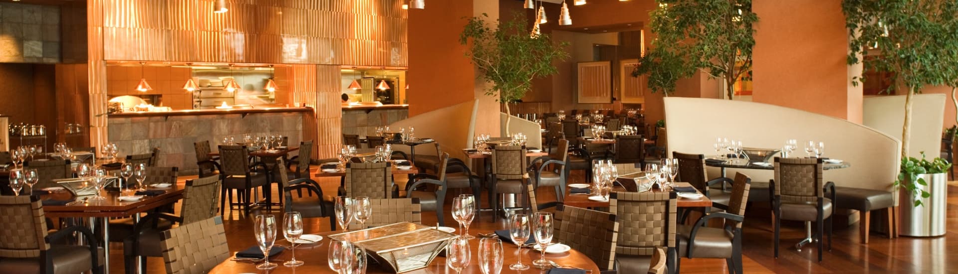 An elegant restaurant dining room with tables set with wine glasses, small plates, napkins and silverware, and an open pass through area with lamps ready for plates to be set to take to diners.