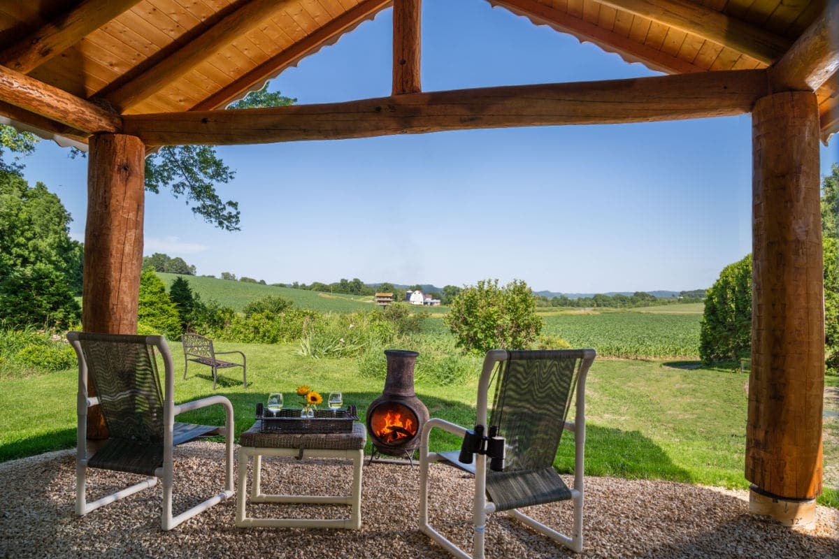A cozy porch with two chairs, a fire pit, and a scenic view of lush greenery and distant hills.