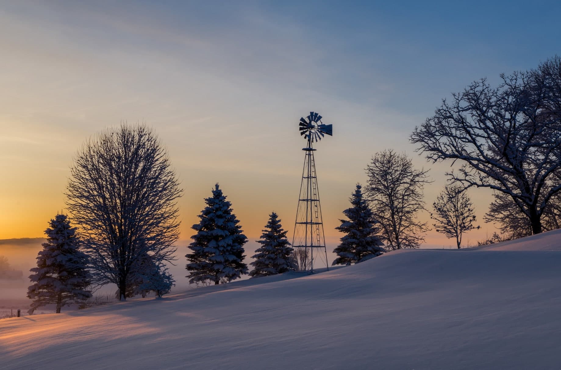 A silhouetted windmill stands among snow-covered trees at sunset.