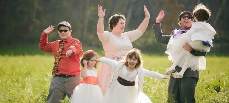A joyful group of five people, including children in white dresses, celebrates outdoors with playful poses.