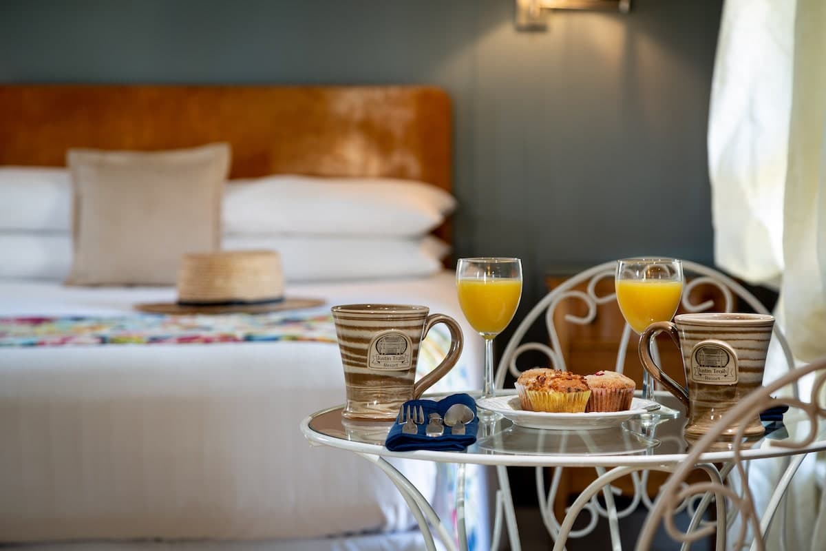 A cozy bedroom setting with two mugs, orange juice glasses, and pastries on a small table in front of a neatly made bed.