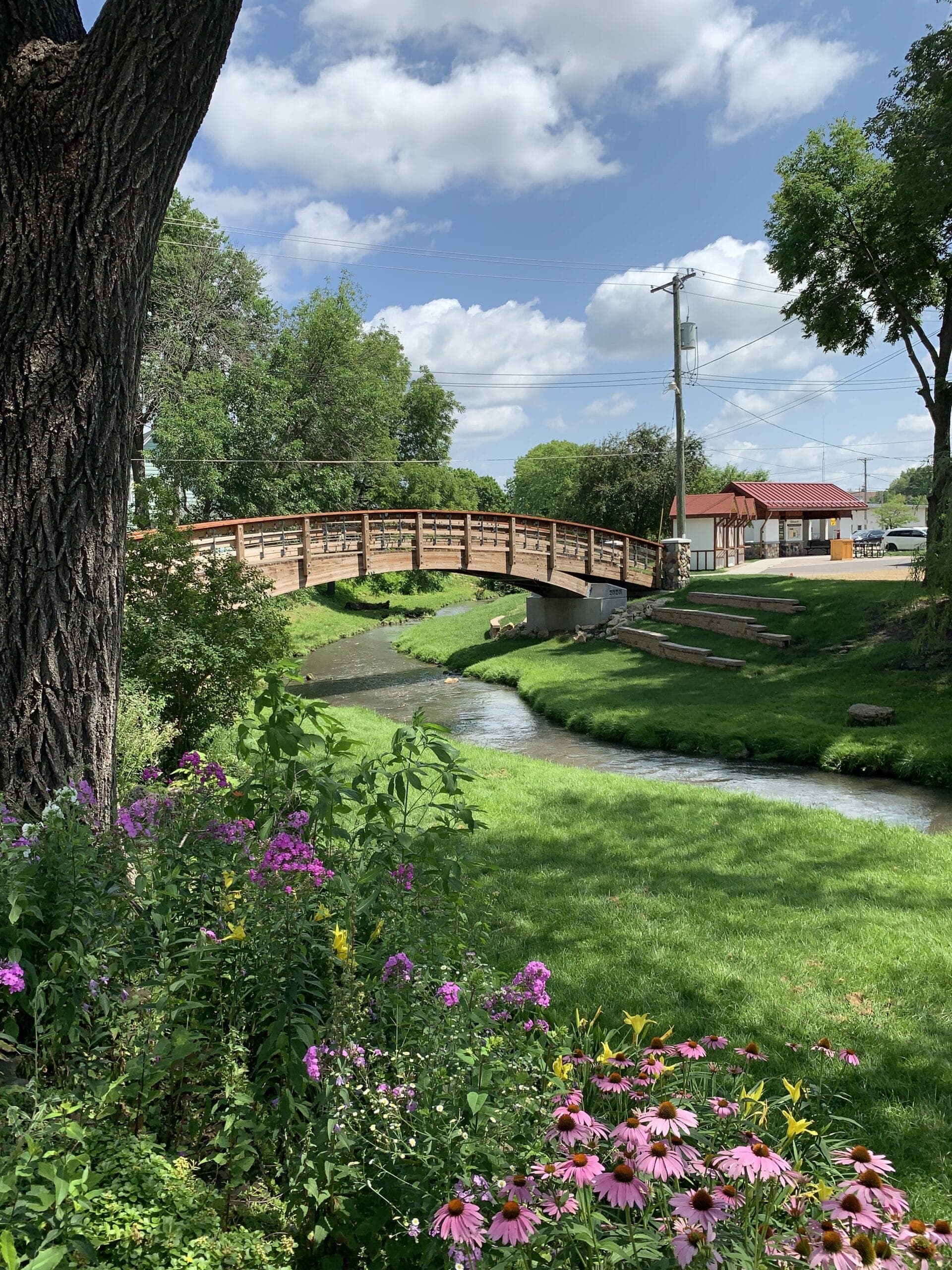 A wooden bridge spans a small creek surrounded by greenery and colorful flowers under a partly cloudy sky.