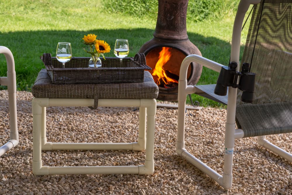Left Image Alt Text
A small table outside with 2 glasses of white wine, a vase of sunflowers, chairs on either side, and a fire in a firepit in front of the table and chairs.