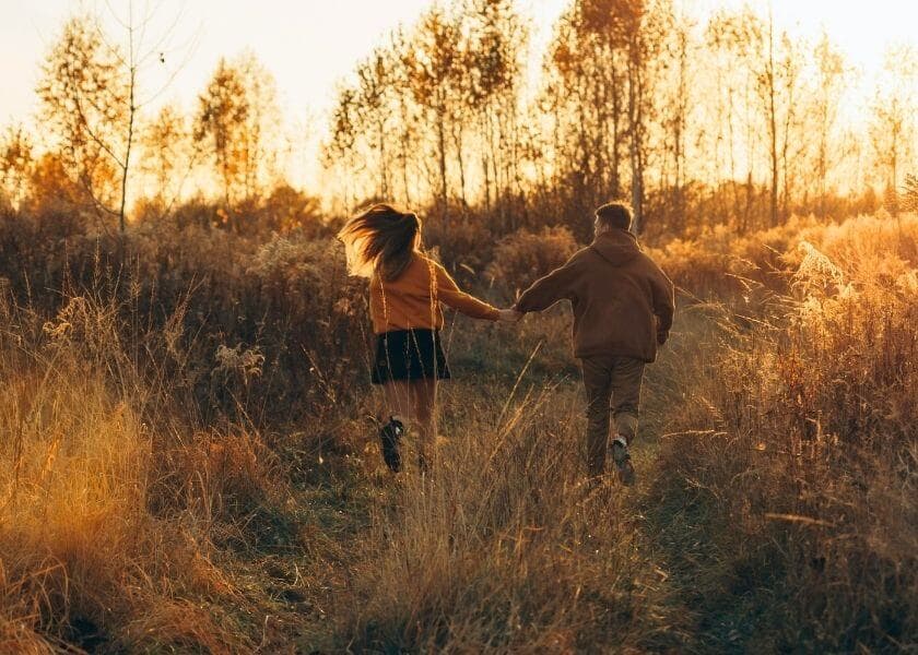 A couple walks hand-in-hand down a sunlit, grassy path surrounded by autumn foliage.