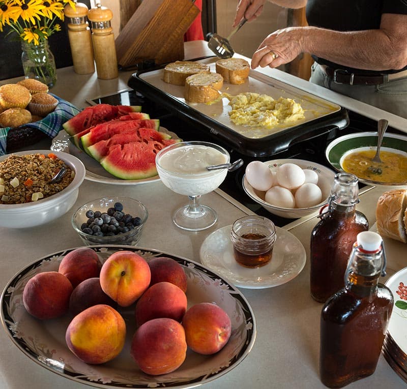 A counter with breakfast foods of scrambled eggs, buttered bread, granola, yogurt, fresh berries, slices of watermelon, a bowl of peaches, hard boiled eggs, and bottles of real maple syrup.