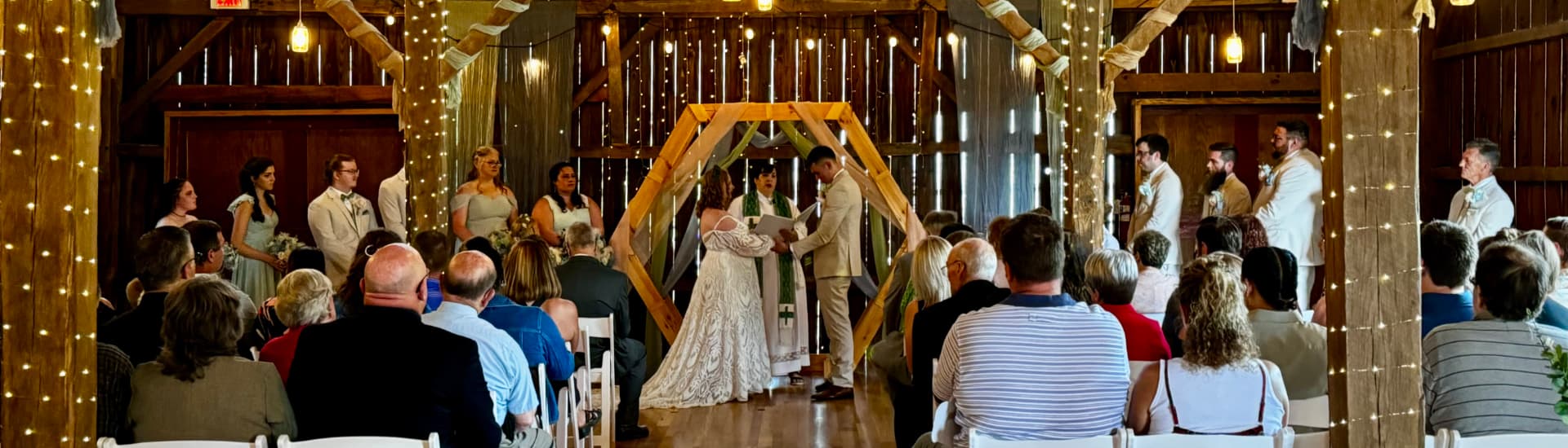 A couple dressed in wetting attire saying their vows in an elegantly decorated barn with bridesmaids and groomsmen on either side of them, and guests sitting in chairs watching them.