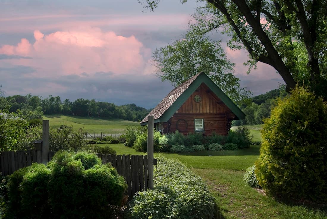 A quaint log cabin with a green roof nestled in a lush garden against a dramatic sky.